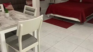 Ein Zimmer mit einem Bett, einem Esstisch mit Stühlen, einem kleinen Tisch mit Pflanze und einem Bild an der Wand.