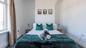 Un lit bien fait, recouvert d’une couverture verte et agrémenté d’oreillers, une fenêtre avec des rideaux et trois tableaux encadrés accrochés au mur.
