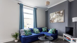 Un salon moderne avec un canapé bleu, une table basse ronde et un téléviseur à écran plat posé sur un meuble blanc.