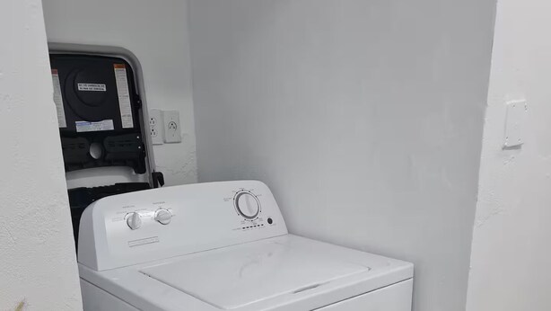 A compact, white front-loading washer and dryer unit in a corner of a room with a tiled floor and a small potted plant. - The Castle ᴴ (Philipsburg)