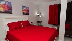 Une chambre avec une couvre-lit rouge, une tête de lit blanche, deux tableaux accrochés au mur, une table de chevet avec une lampe et un rideau rouge.