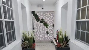 Un couloir aux murs blancs, orné de deux grands pots de plantes, d’un mur décoratif et d’un luminaire au plafond.