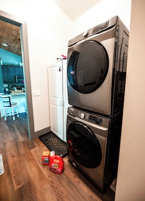 A laundry area with stacked washers and dryers, a white door, and a kitchen visible in the background.