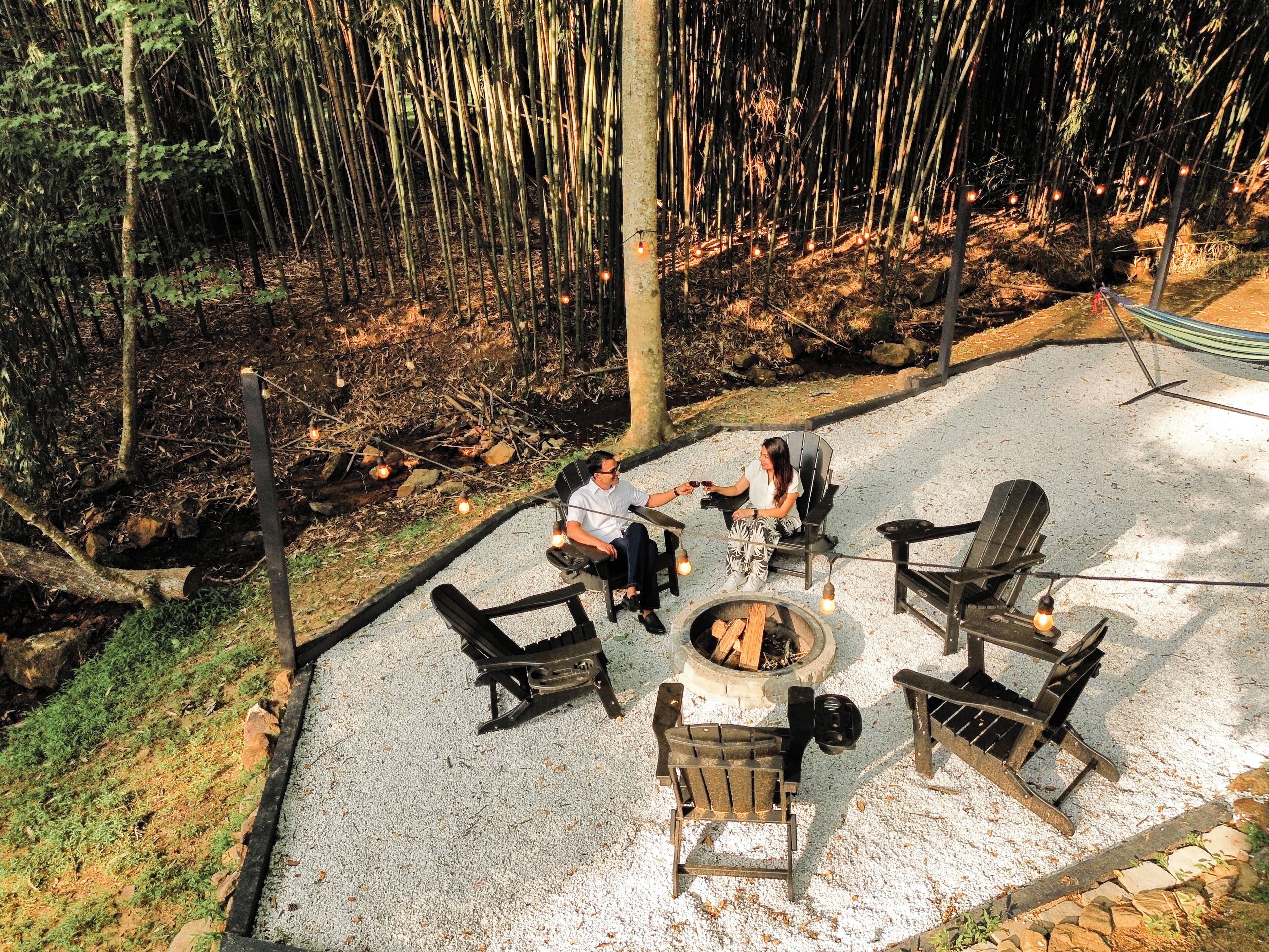 An outdoor seating area with a fire pit, surrounded by chairs and tables, set in a forested environment.