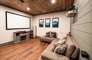 Une salle de cinéma maison dotée d’un grand écran, d’un plancher en bois, d’un canapé gris et de décorations murales sur le thème de la montagne.