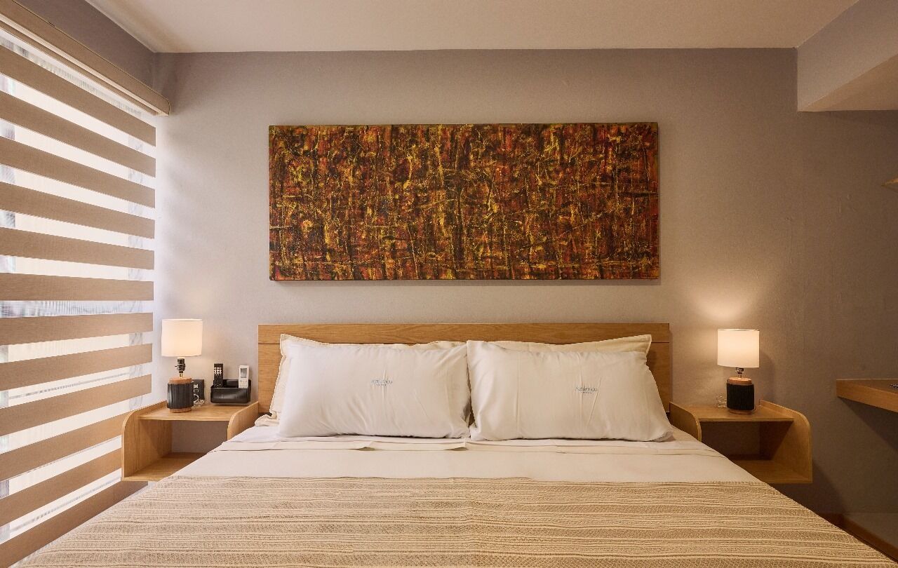 A hotel room with a large bed, two bedside tables with lamps, a wall-mounted abstract painting, and a window with blinds.
