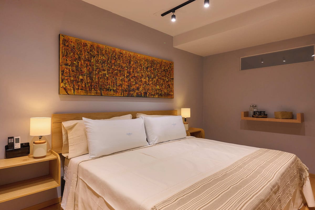 A neatly made bed with white linens, two bedside tables with lamps, a large abstract painting on the wall, and a small shelf with decorative items.