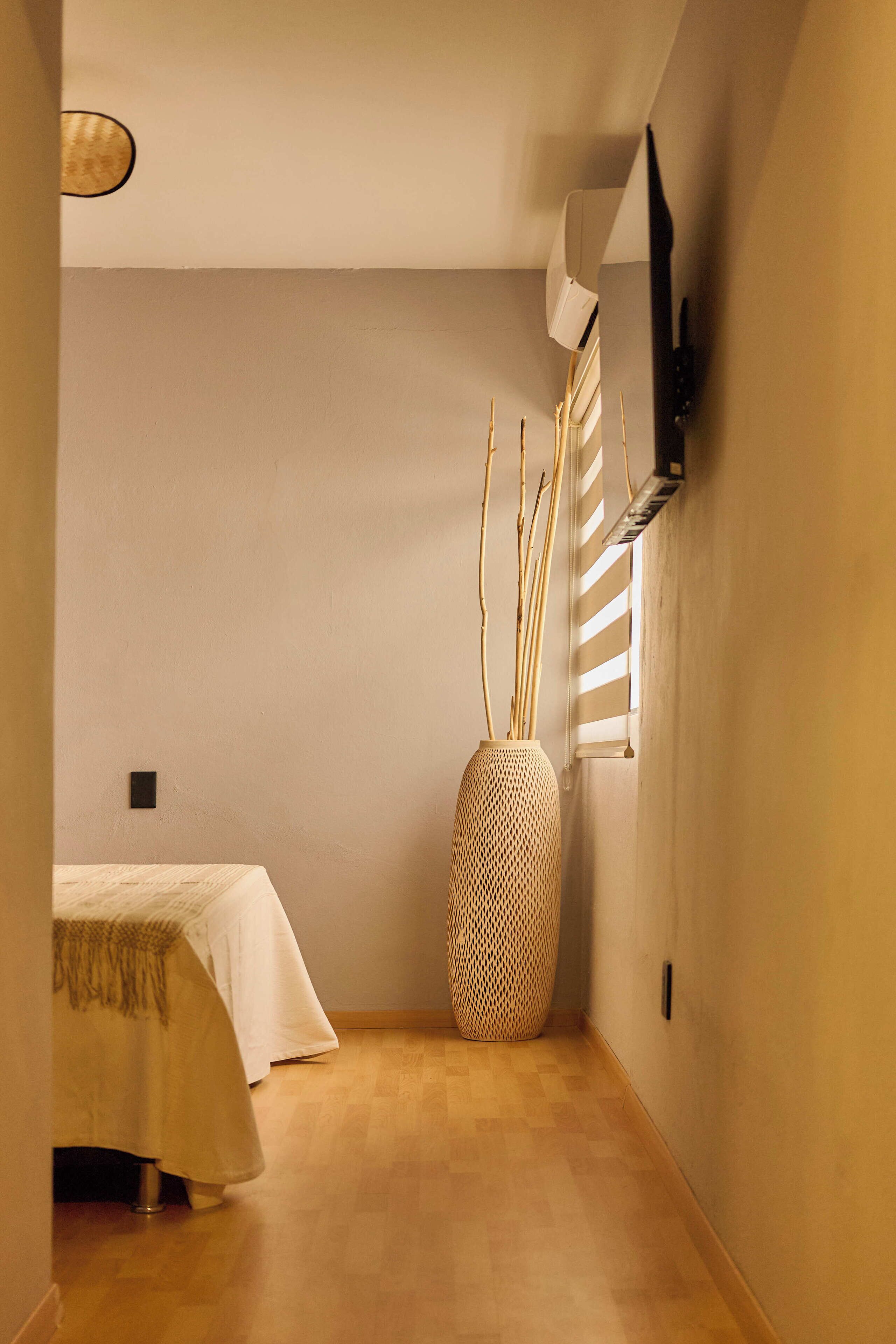 Un pasillo estrecho con una cama a la izquierda, un jarrón grande tejido y una ventana con persianas.