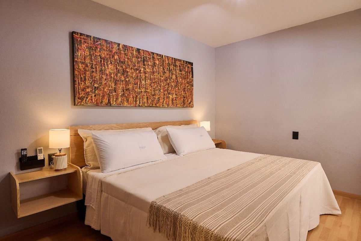 A hotel room with a large abstract painting on the wall, a bed with white linens, a bedside table with a lamp, and a wooden nightstand.
