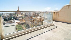 A balcony with a glass railing overlooking a cityscape with historic buildings and a church spire. - Ghajnsielem Retreat 4 Bedrooms, Panoramic Terrace (Ghajnsielem)