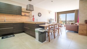 A modern kitchen with a central island, a dining area with chairs, a sofa, and a large window offering a city view. - Ghajnsielem Retreat 4 Bedrooms, Panoramic Terrace (Ghajnsielem)