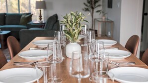 Een eettafel gedekt met witte borden, glazen en bestek, met in het midden een witte vaas met groene bladeren.