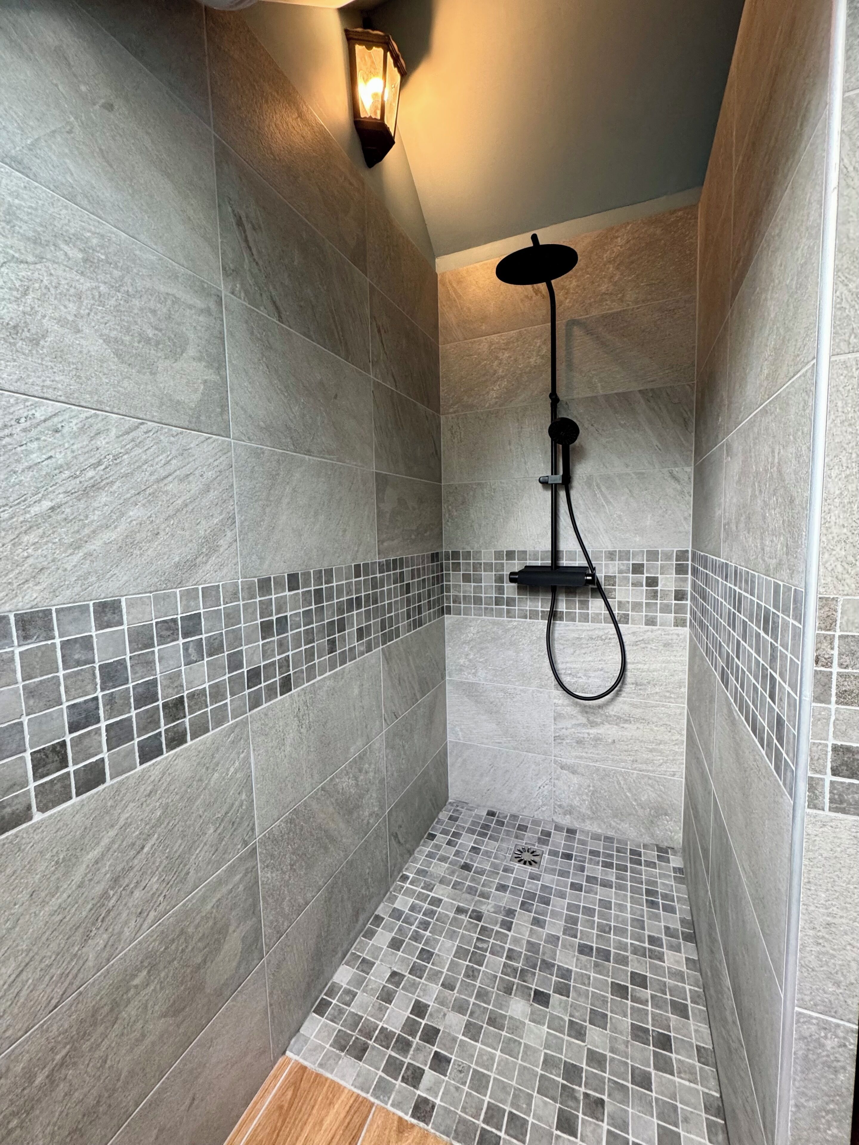 Une douche avec un sol et des murs carrelés, une douchette à main et un luminaire fixé au mur.