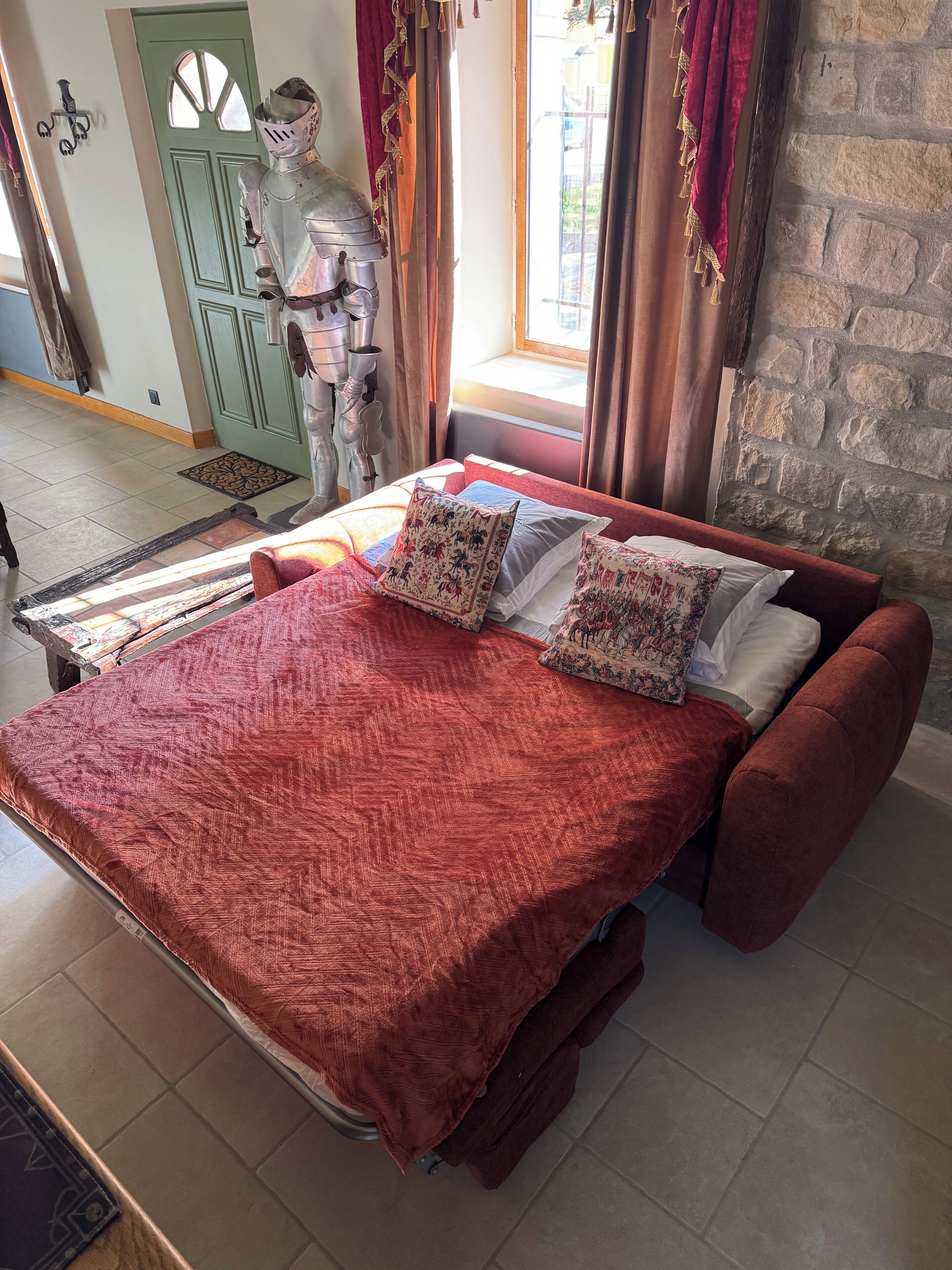 A bed with a red quilt and patterned pillows, a stone wall, and a green door.