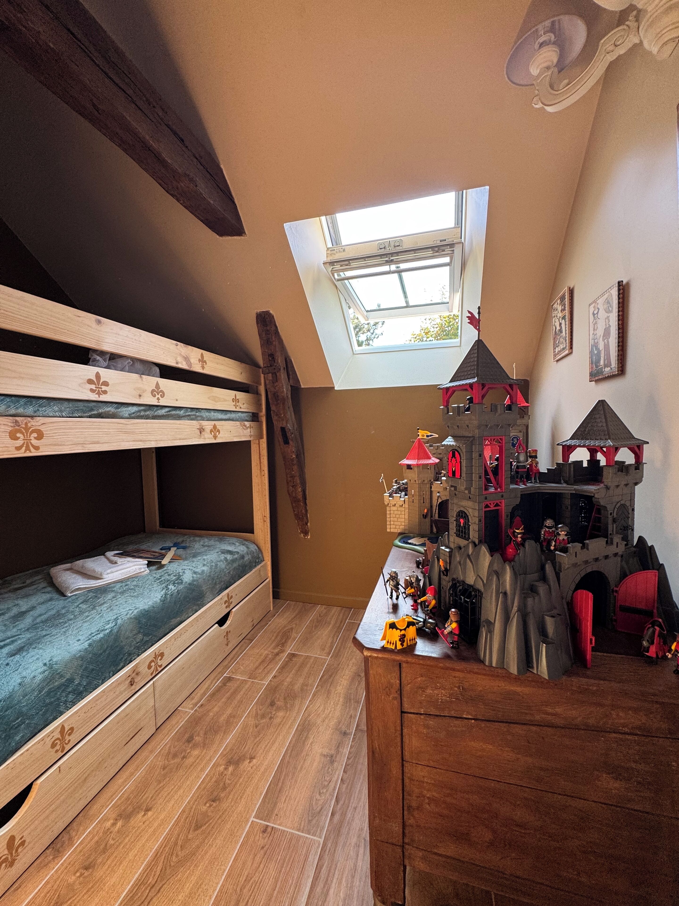Ein Zimmer mit einem Etagenbett, einem Holztisch mit einem Burgmodell und einem Oberlicht.