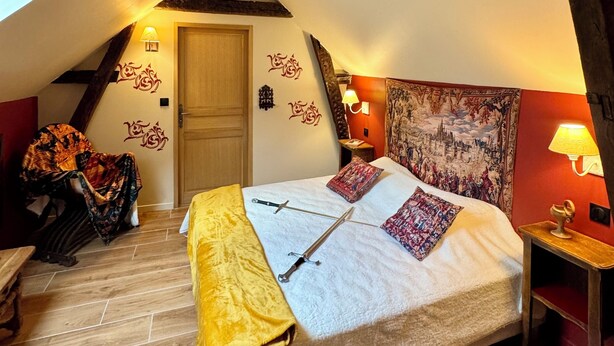 A bedroom with a sloped ceiling, exposed wooden beams, a bed with a yellow throw, patterned pillows, and a tapestry on the wall. - Unusual: The Knights' Lodge—Medieval-themed, air-conditioned, and featuring balneotherapy (Sens)