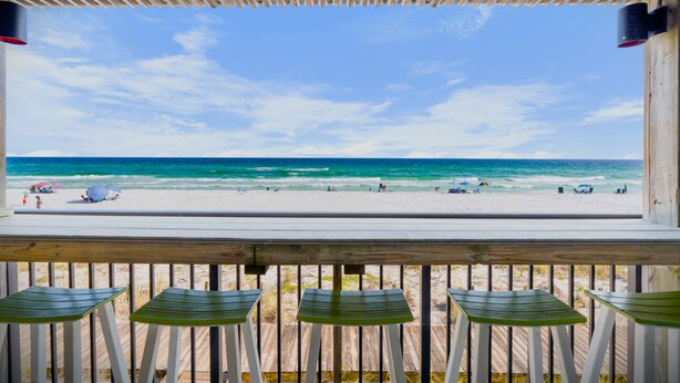 Blick auf einen Sandstrand mit Menschen und Sonnenschirmen von einer hölzernen Terrasse mit Geländer.