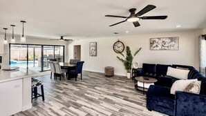 A spacious living area with a dark blue sofa, a dining table with chairs, and a kitchen area with a white countertop and pendant lights.