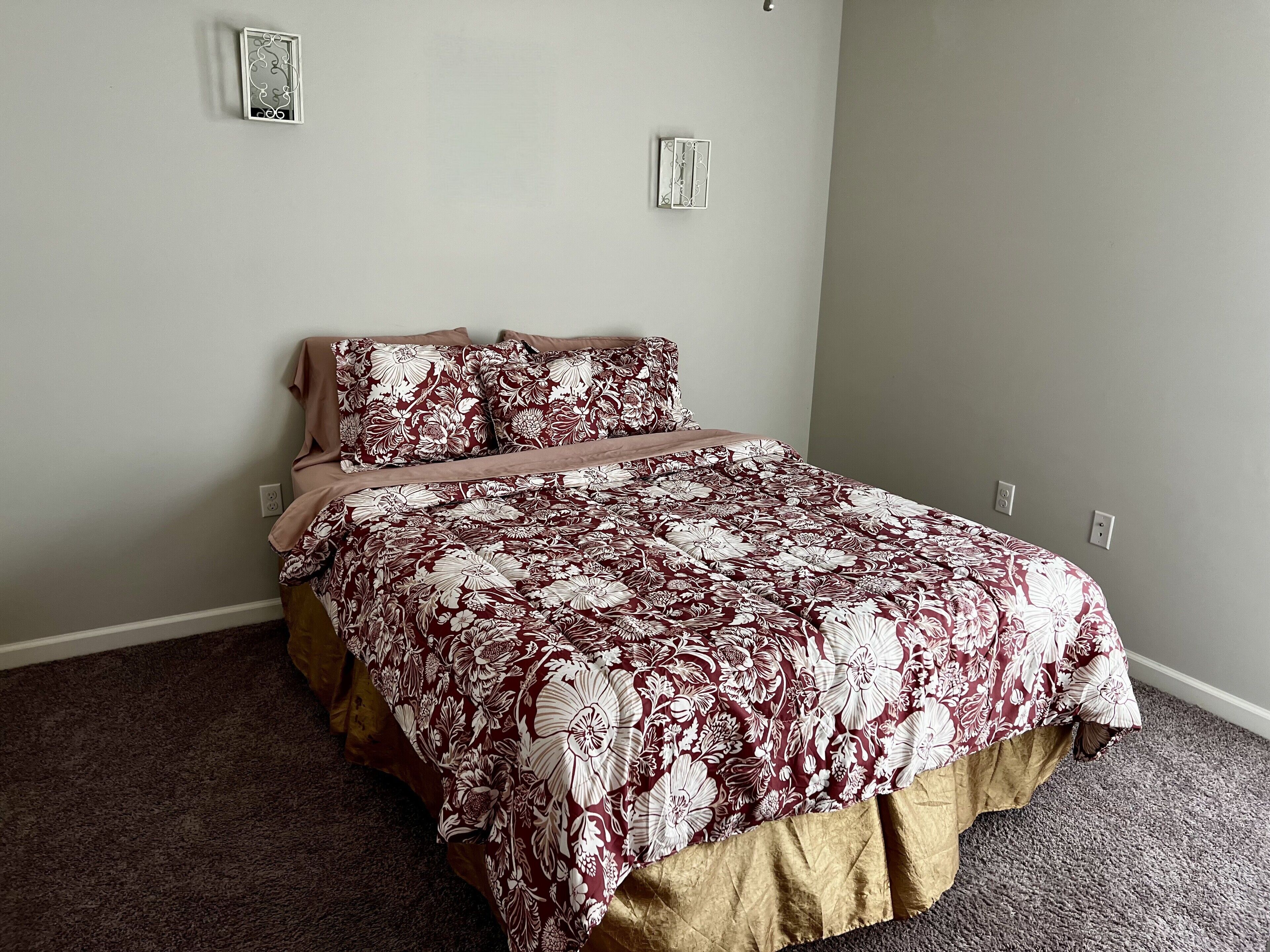 A bedroom with a floral bedspread, a beige bed skirt, and two wall-mounted light switches.