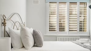 Un letto rifatto con lenzuola bianche, una lampada da comodino e una finestra con persiane.
