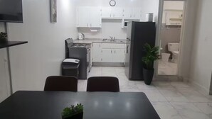 Una cocina moderna con armarios blancos, horno integrado y refrigerador. Hay una mesa de comedor con sillas y una pequeña planta en maceta encima.