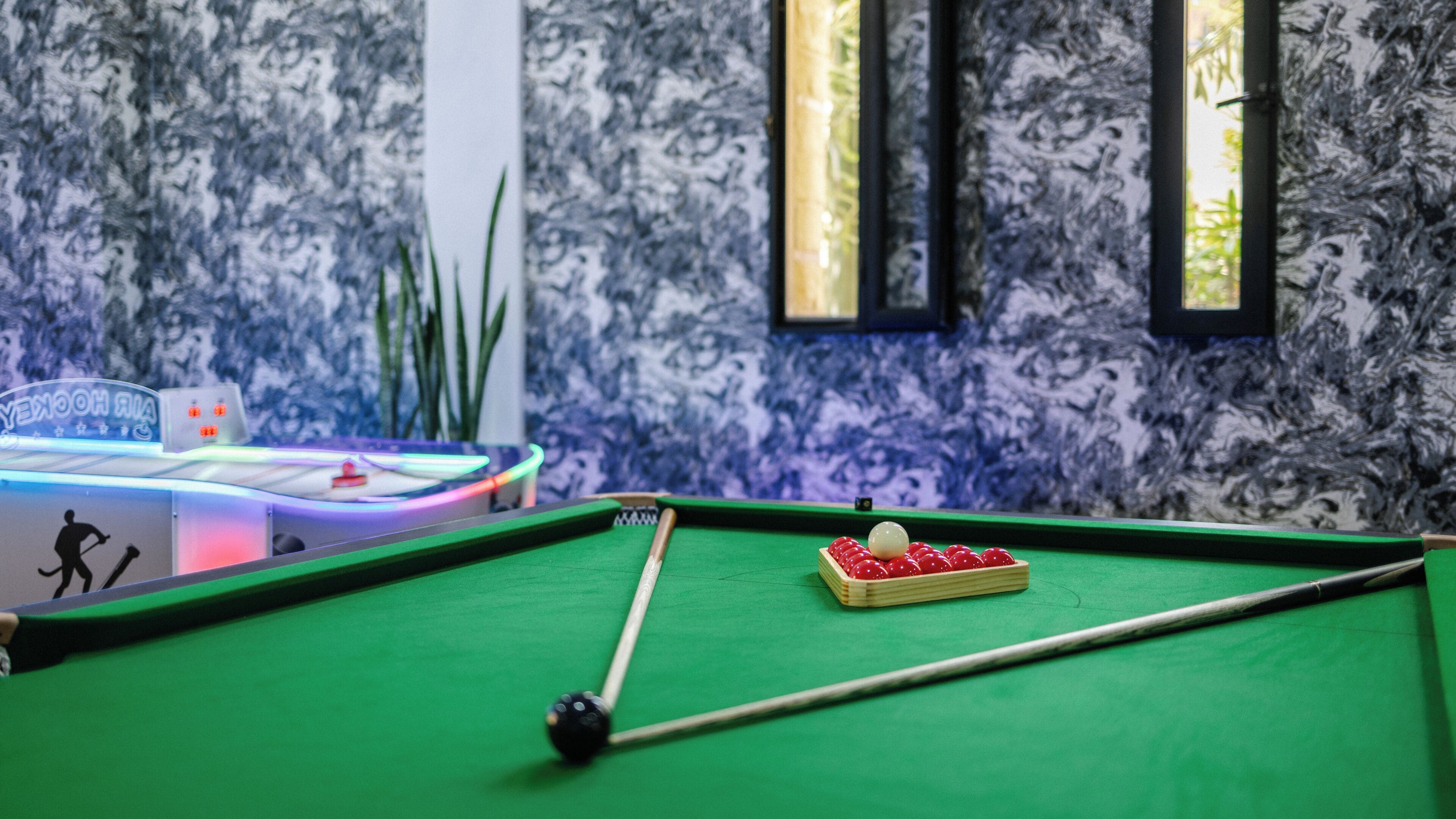 Ein Billardtisch mit Spielzeugkeule und Kugeln, ein neonbeleuchteter Spielautomat und eine Wand mit blau-weißem Muster.