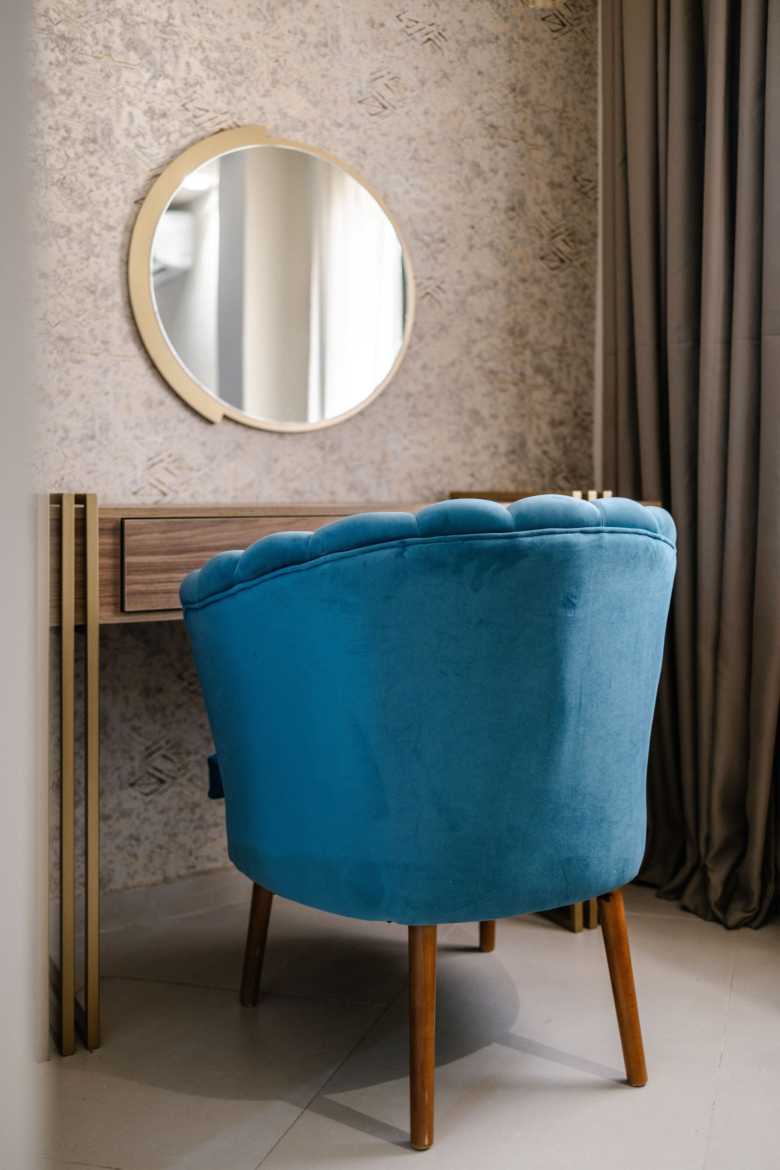 A blue chair with wooden legs and a round mirror on a wall with a textured pattern.