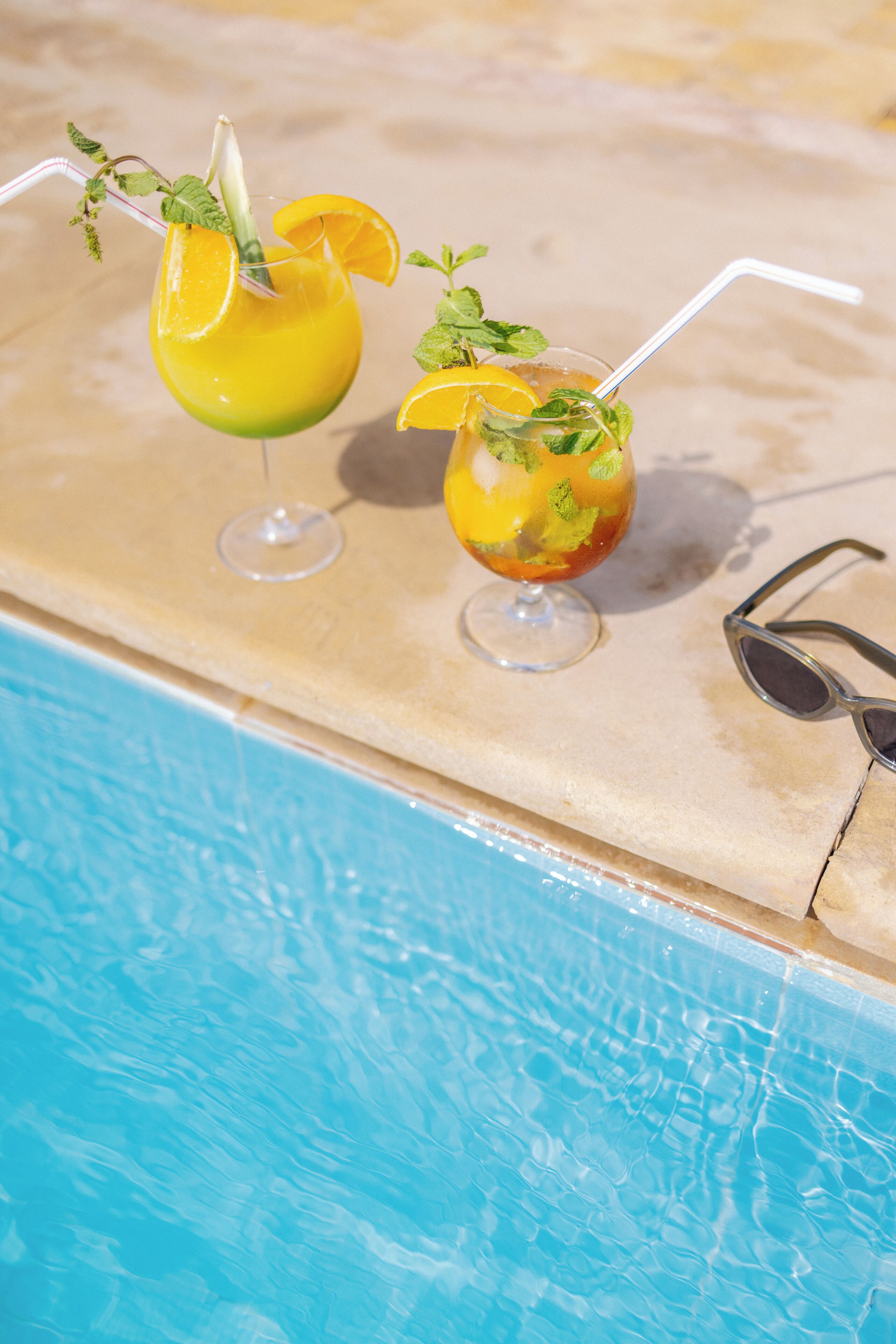 Two cocktails with straws and garnishes on a poolside table next to a pool.