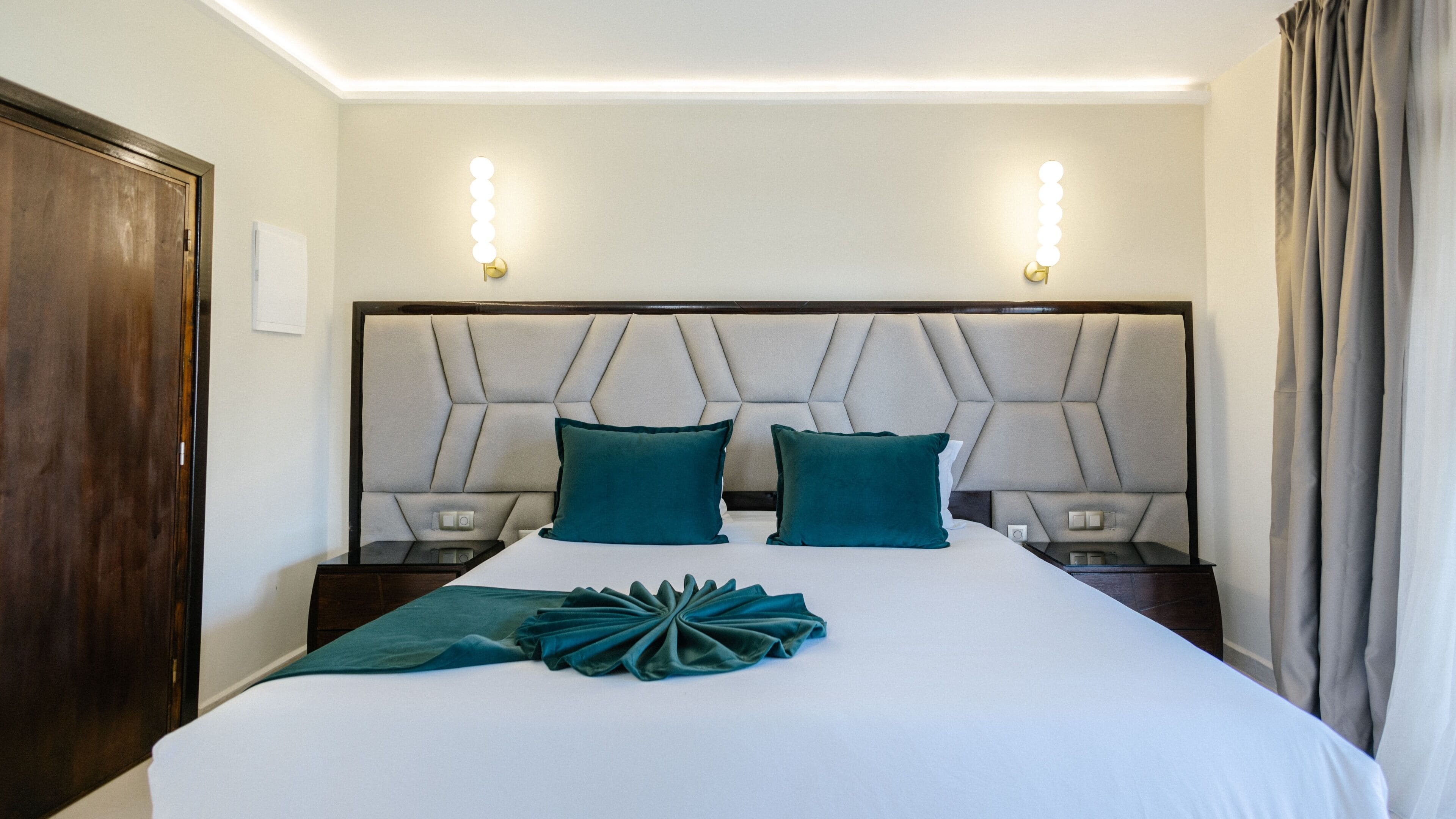 Habitación de hotel con una cama grande, dos mesitas de noche, un cabecero moderno y una lámpara fijada a la pared.