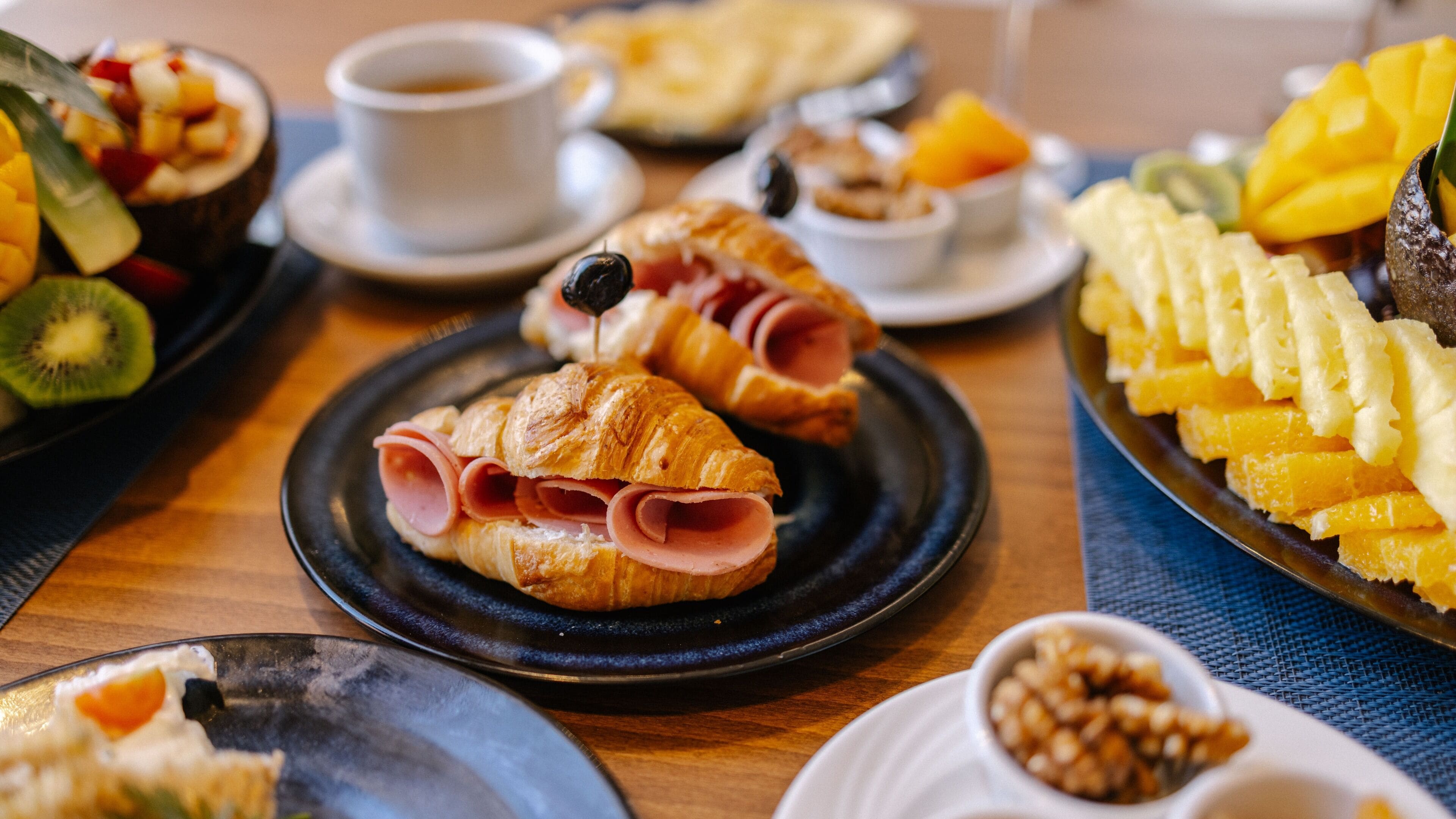 Een ontbijt met croissants, ham, fruit en koffie op een houten tafel.