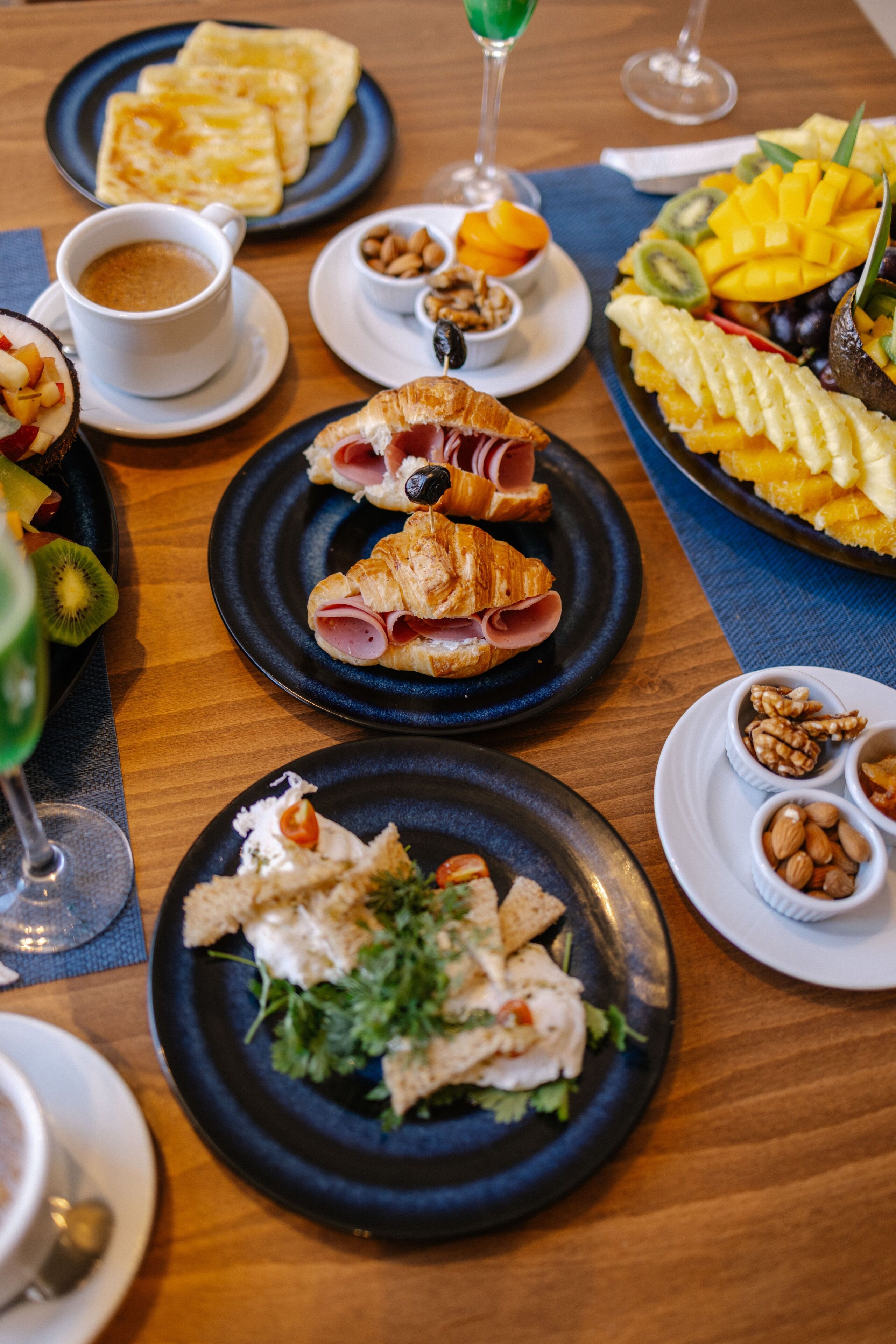 Una mesa dispuesta con varios platos que incluyen cruasanes, fruta y frutos secos, junto con tazas de café y un vaso de bebida verde.