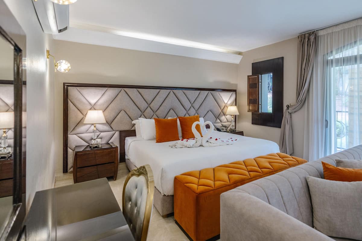 A hotel room with a large bed, a sofa, a nightstand, a lamp, and a window with curtains.