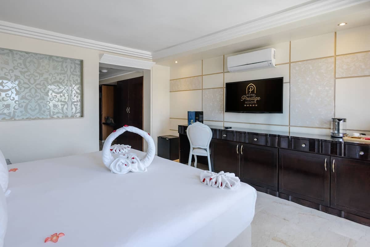 A hotel room with a large bed, a TV on the wall, a minibar, and a view of the hallway.