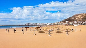 A beach with sun loungers and umbrellas, a clear sky, and buildings in the background. - Hotel Prestige Agadir (Agadir)