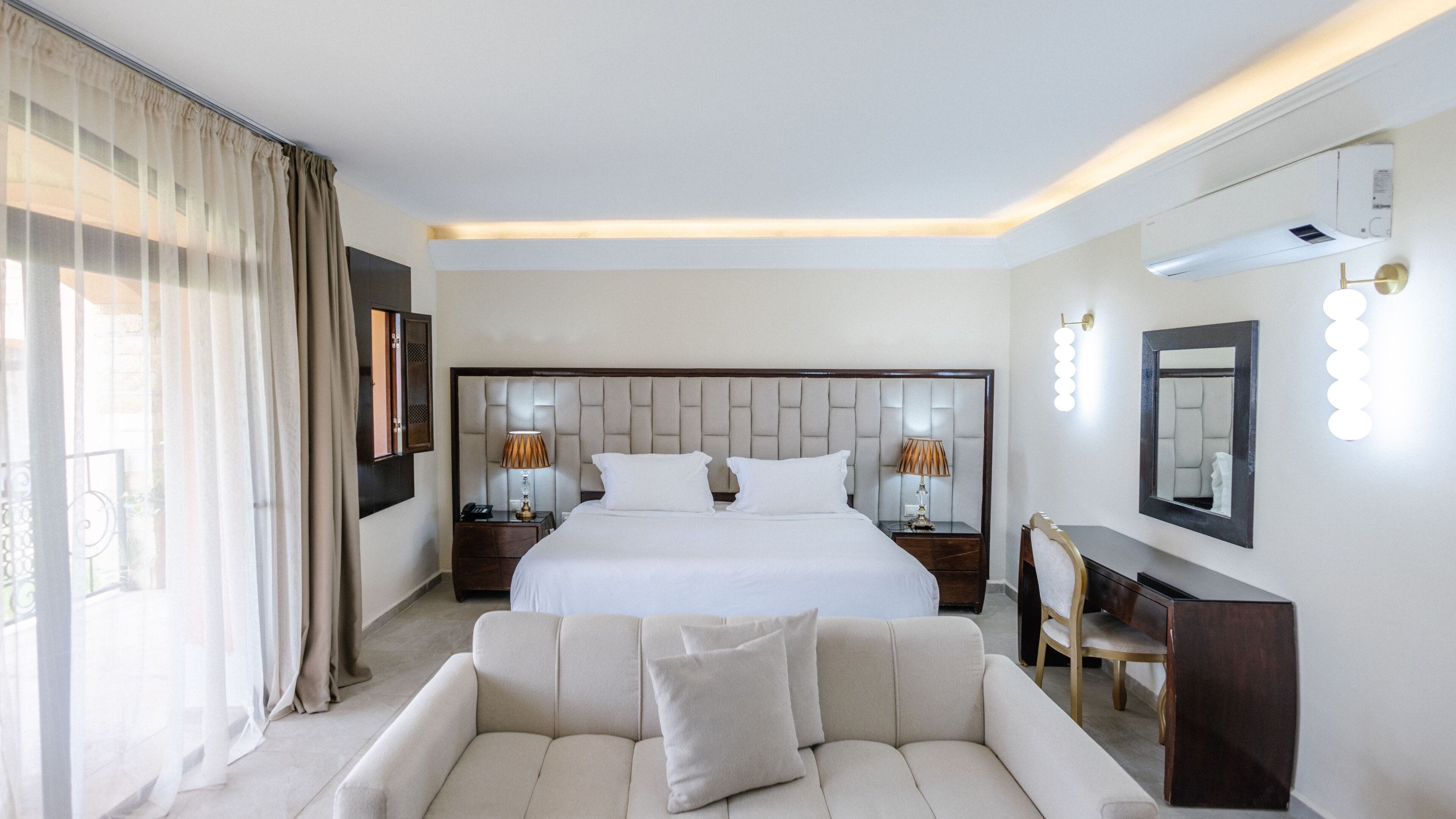 A modern hotel room with a large bed, two bedside tables with lamps, a sofa, a mirror with lights, and a view of the outdoors.