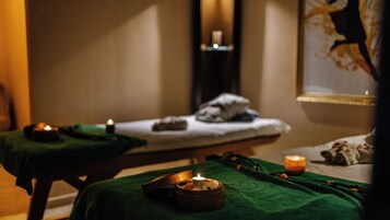 Un cuarto de spa con dos camillas cubiertas con toallas verdes, velas y un pequeño cuenco con una vela en una de las camillas.