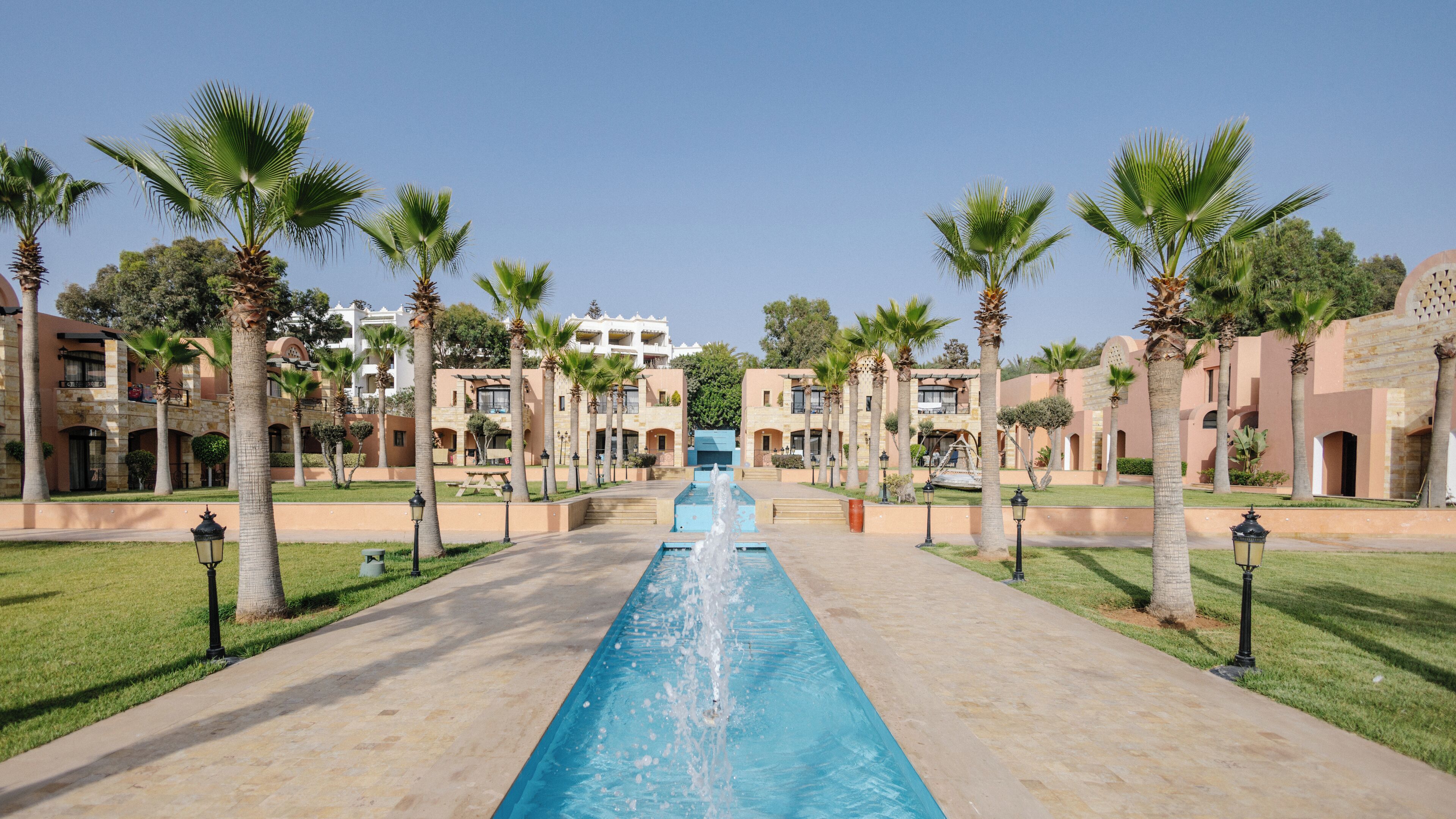 Ein Pool mit Wasserspiel in der Mitte, umgeben von Palmen und Gebäuden mit Balkonen.