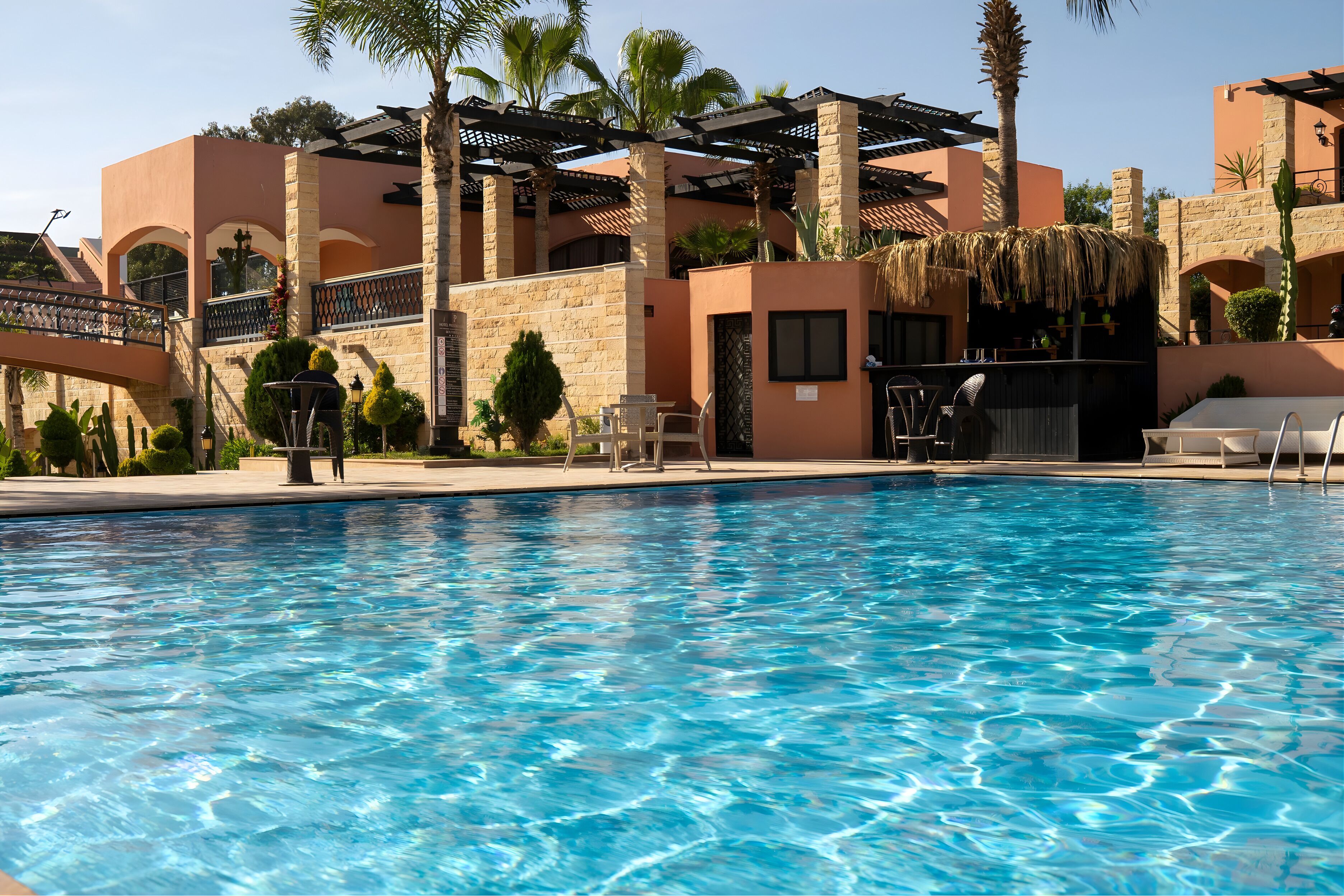 A swimming pool with lounge chairs and a bar area, surrounded by palm trees and a building with arches and a thatched roof.