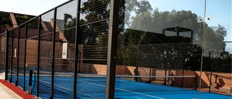 Una cancha de tenis con una red, rodeada por una valla, y un camino con alfombra roja.