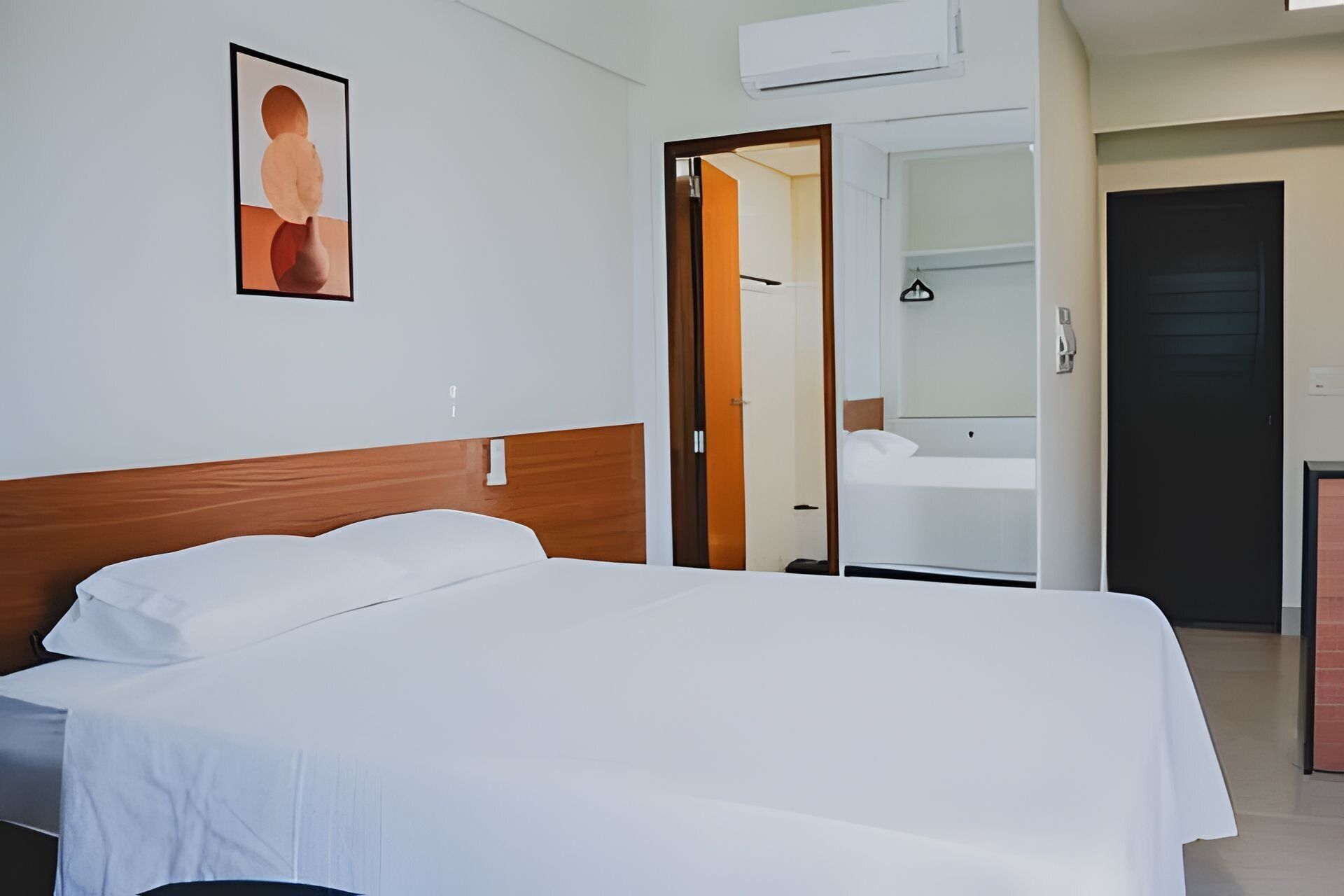 A hotel room with a bed, a wooden headboard, a white wall, and a door leading to a bathroom.