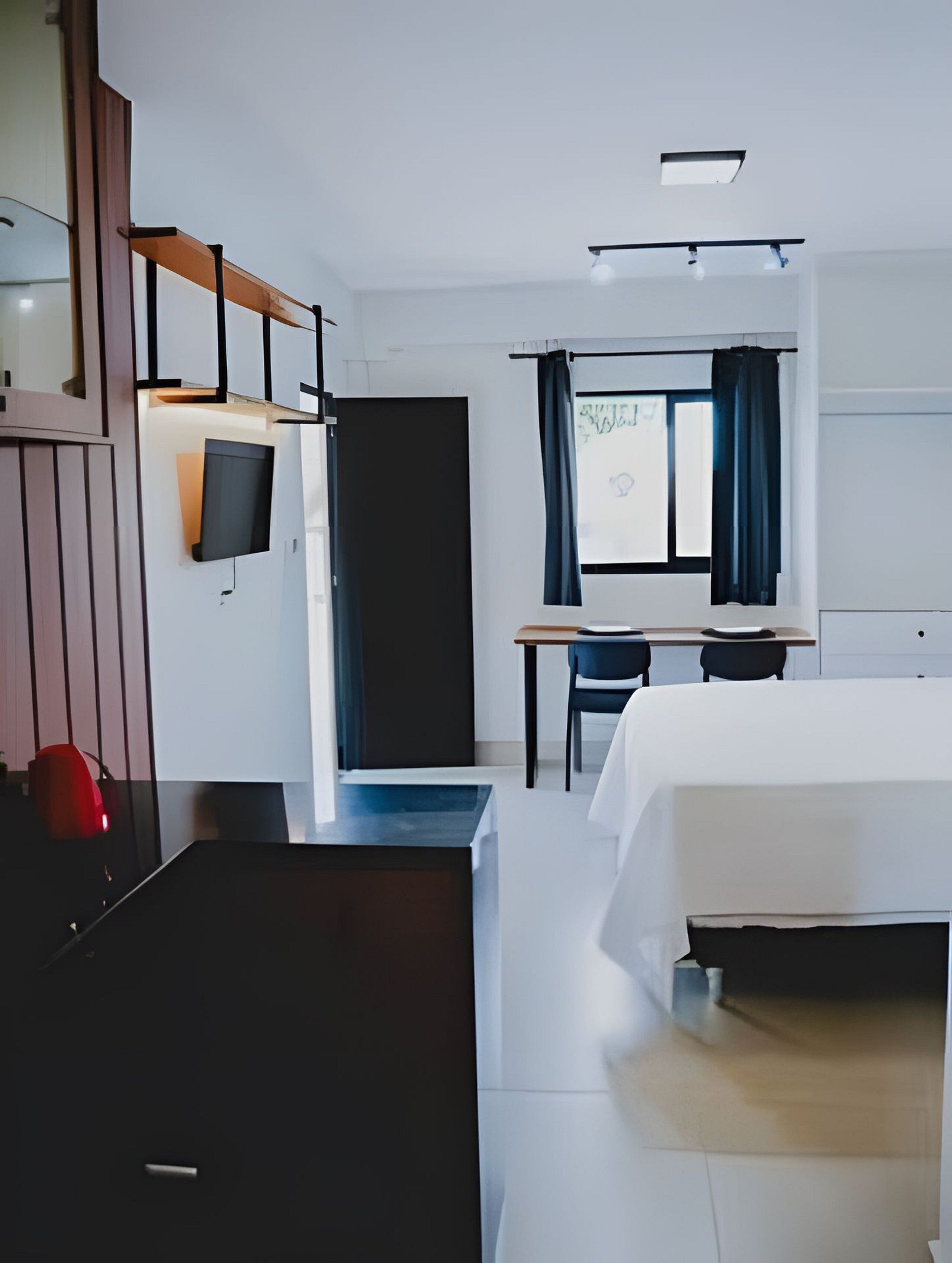 A hotel room with a bed, a desk, a chair, a TV mounted on the wall, and a window with curtains.