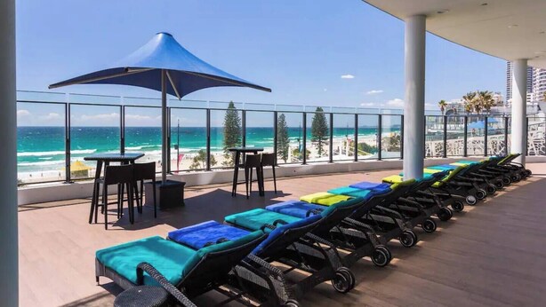 A balcony with lounge chairs and a table, overlooking a beach and ocean. - Absolute beachfront luxury in Surfers Paradise — a designer 3-bedroom apartment on level 11 of the iconic Peppers Soul tower. Panoramic ocean views, wraparound balcony, resort pools, spa & gym. Sleeps 6. Steps to the sand, 300m to Cavill Ave. (Surfers Paradise)