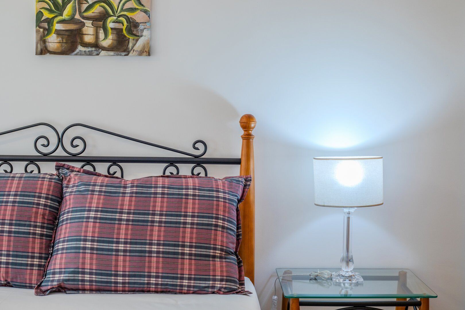 A bed with plaid pillows, a metal headboard, a wooden post, a glass table with a lamp, and a painting of plants on the wall.