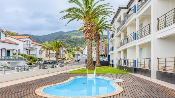 Zona de piscina con terraza de madera, palmeras y vista a una calle residencial con edificios y un paisaje montañoso al fondo.