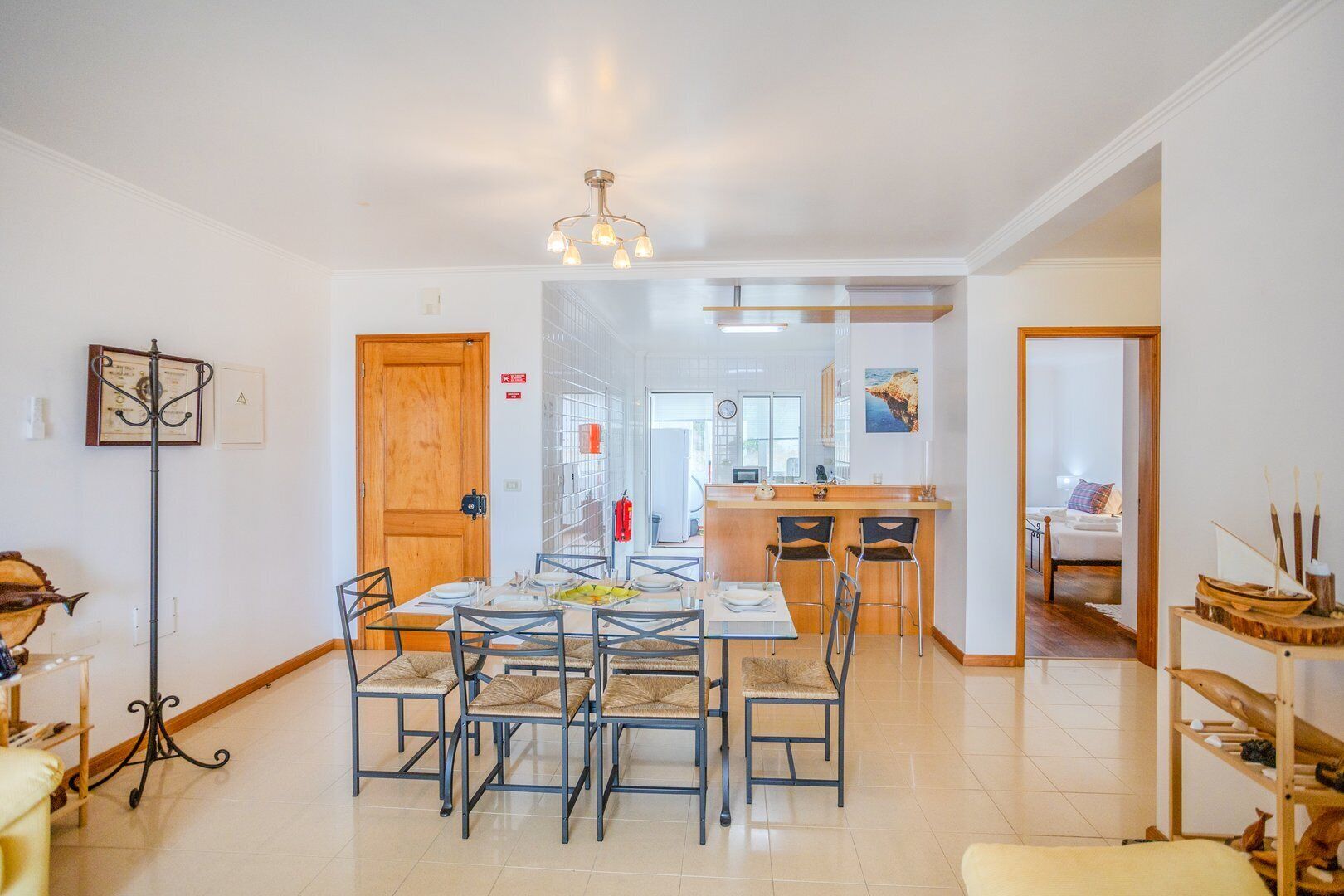 A dining area with a table set for six, a kitchen in the background, and a bedroom visible through an open door.
