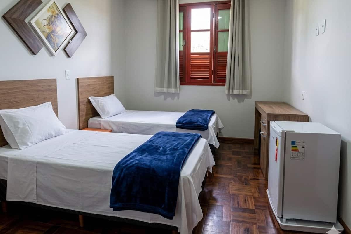 A hotel room with two beds, a wooden floor, a window with curtains, and a small refrigerator.