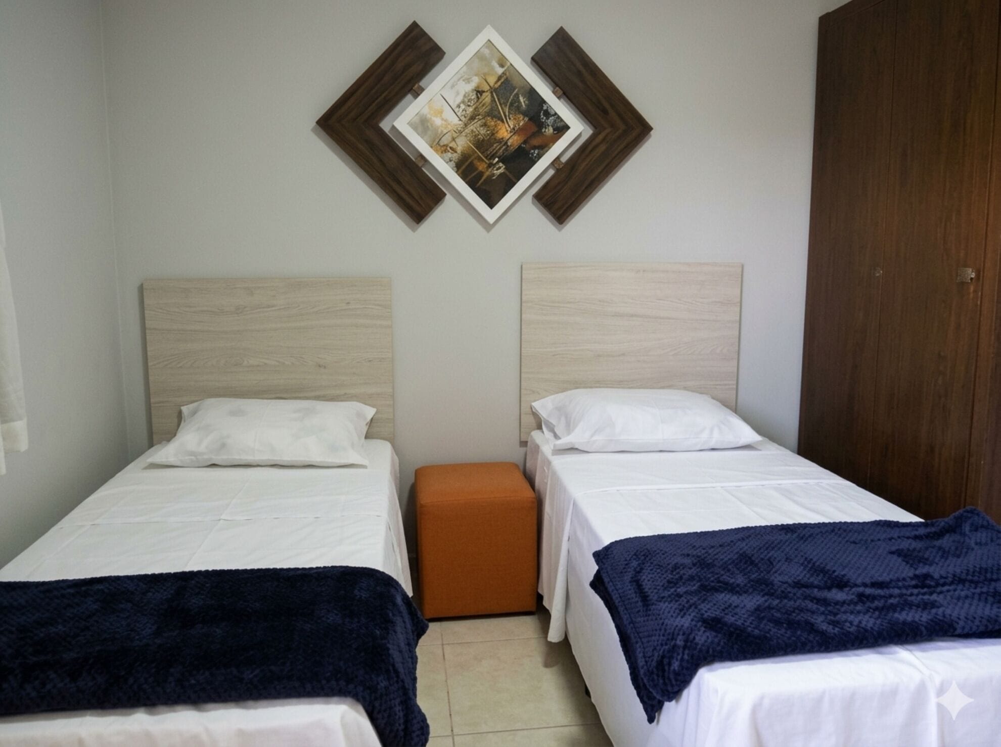Quarto com duas camas de solteiro, um banquinho laranja pequeno e um guarda-roupa de madeira.