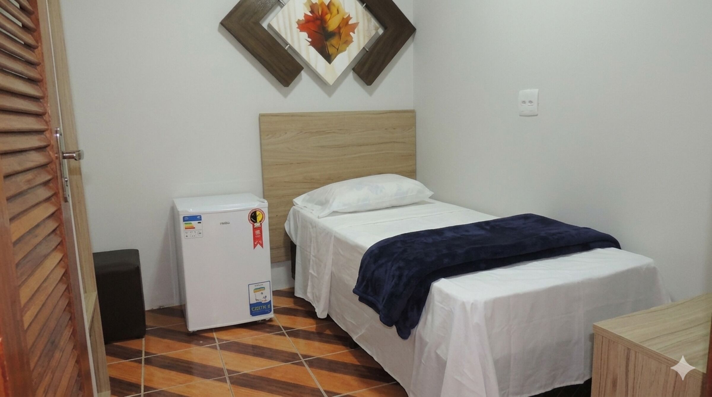 A small, clean room with a bed, a nightstand, a refrigerator, and a wall-mounted picture.