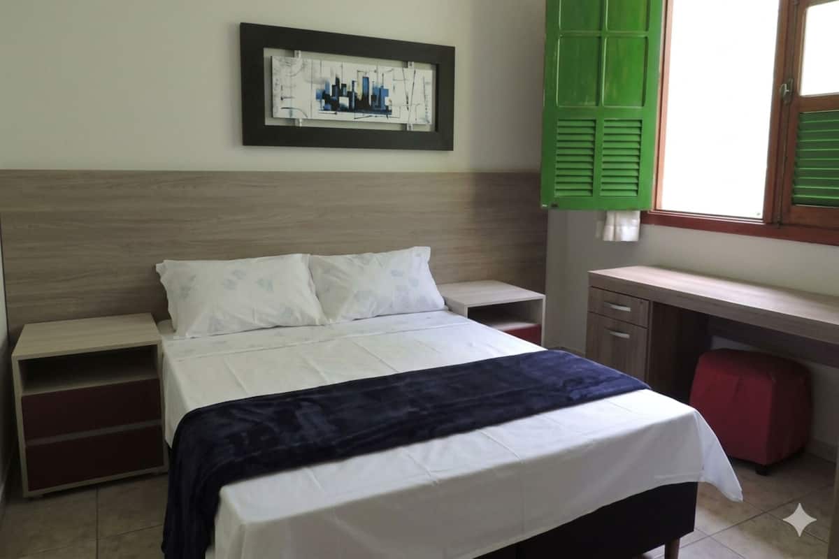 A bedroom with a bed, a nightstand, a dresser, a desk, and a window with green shutters.