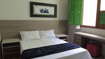 Un dormitorio con cama, mesita de noche, cómoda, escritorio y ventana con contraventanas verdes.